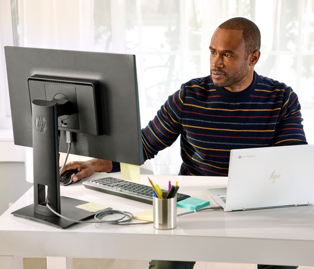 Una persona con una camisa a rayas sentada en un escritorio trabaja en un Chromebook conectado a un monitor externo.
