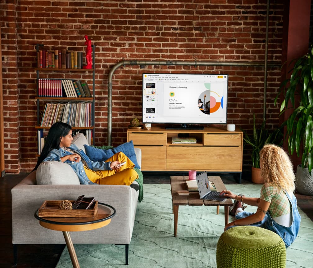 Una persona sentada en un sofá viendo una presentación en la televisión. Otra persona está sentada en el suelo junto a una mesa de centro con un Chromebook.