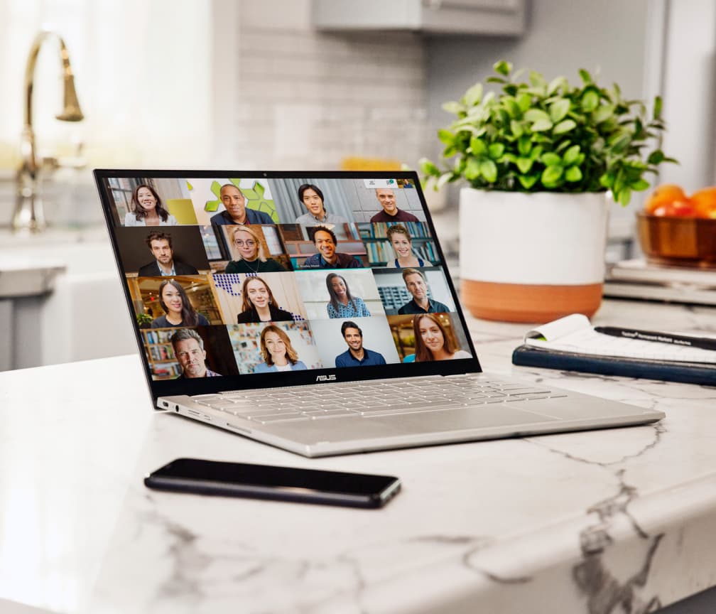 Hay un Chromebook abierto sobre un mostrador de mármol blanco, con un teléfono móvil a la izquierda y una planta al fondo.